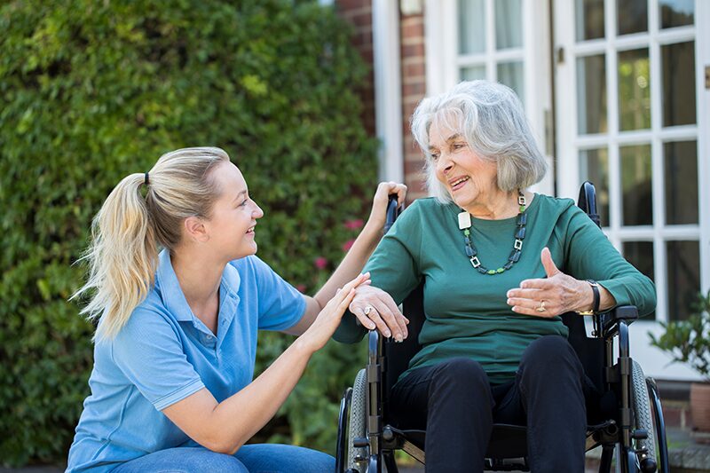 Une Femme Âgée En Fauteuil roulant à l'extérieur de la maison accompagnée d'une jeune femme