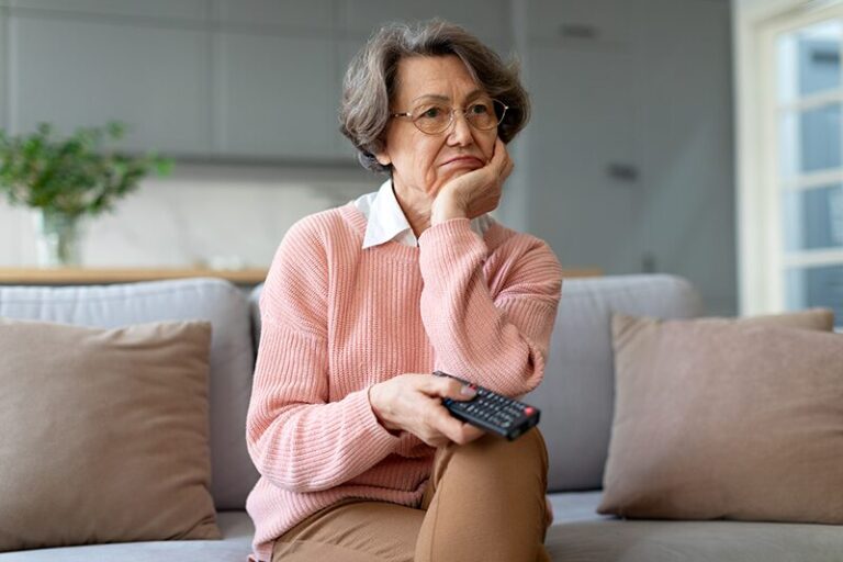 Femme senior assise sur un canapé, l'air préoccupé, tenant une télécommande, illustrant l'incertitude face au chômage et aux nouvelles lois concernant les seniors.