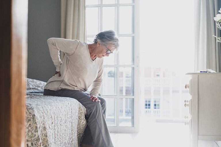 Femme âgée souffrant de douleurs lombaires assise sur un lit, symptôme courant de l'ostéoporose chez les seniors.