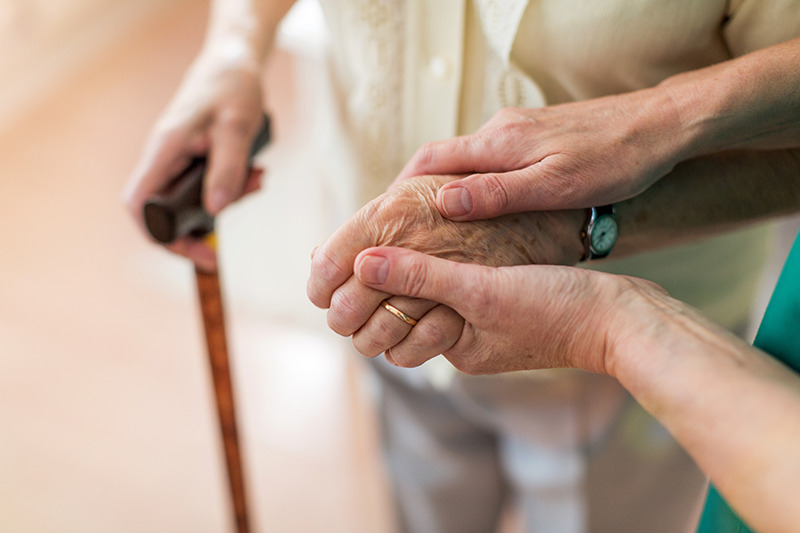 Parkinson héréditaire : une femme âgée atteinte de Parkinson reposant sur une canne à pied et tenant la main d'une infirmière.