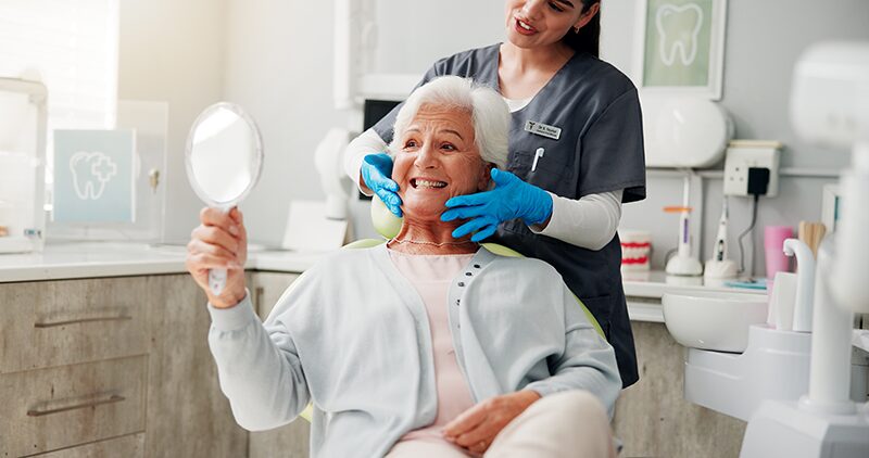 Une personne âgée qui se regarde sa prothèse dentaire en miroir avec la dentiste souriante derrière elle.