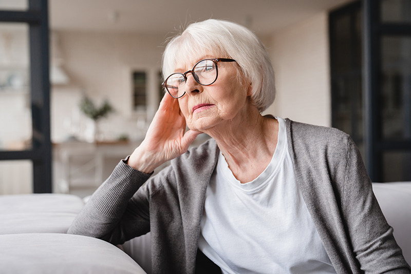 femme âgée d'environ 70 ans, souffrante de dégénérescence cognitive assise toute seule dans le salon de sa maison et réfléchit la main sur le visage.