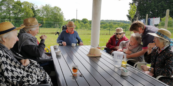 Six personnes âgées prennent leur goûter dehors sur la terrasse accompagné par une assistante de vie