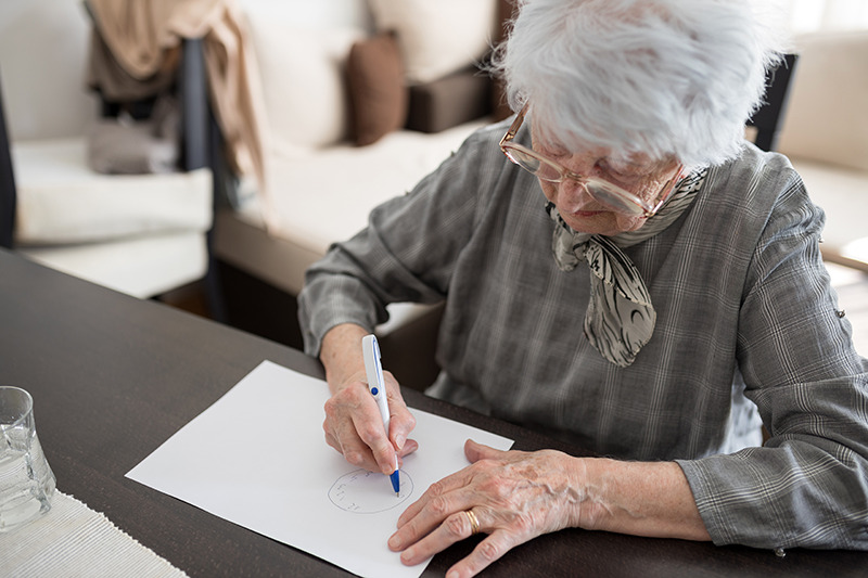 test de l'horloge : une personne âgée, vêtu en gris et portant des lunettes de vue, est entrain de dessiner le cadran d'une horloge sur une feuille blanche.